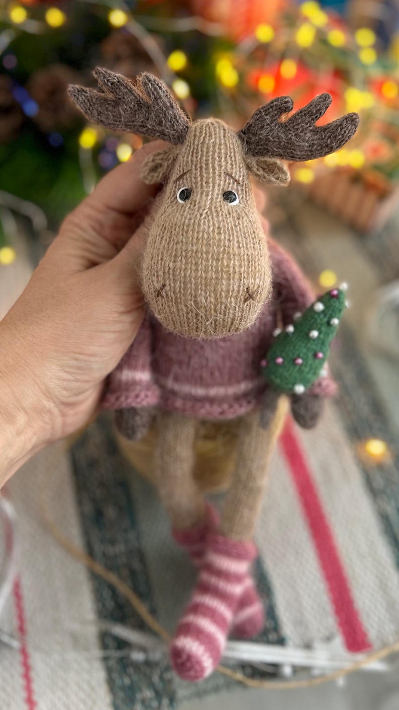 May include: A brown knitted moose toy wearing a pink sweater and holding a small green Christmas tree. The moose has large brown antlers and black eyes.