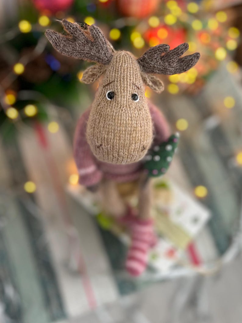 May include: A brown knitted moose toy with a pink sweater and striped socks, holding a small green Christmas tree. The toy is sitting on a striped surface with a blurred background of Christmas lights.