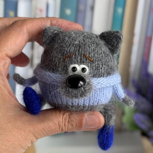 Puede incluir: Una muñeca de gato gris tejida a mano con una bufanda azul y pies azules. El gato tiene ojos negros y una nariz negra. La muñeca se sostiene en una mano, con un fondo borroso de libros y flores moradas.