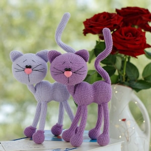 Pode incluir: Dois brinquedos de gato de crochê de cor roxa com narizes cor-de-rosa e olhos pretos. Os gatos estão em pé sobre um livro com um buquê de rosas vermelhas no fundo.