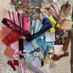 May include: A selection of stationery items, including pens, pencils, highlighters, notebooks, sticky notes, decorative paper clips, and washi tape. The items are in various colours, including pink, blue, and brown. Some items have floral designs.
