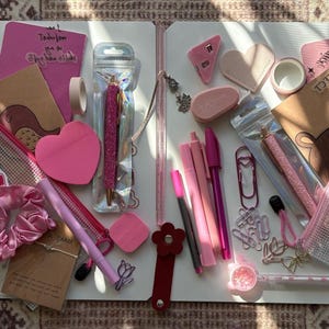 May include: A selection of pink stationery items, including pens, sticky notes, a scrunchie, and a notebook. The items are arranged on a white surface, with various shades of pink and other accessories. The image includes the text "MAKES PERFECT".