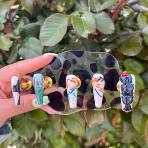 May include: Five different nail designs on a black and white cow print display. The designs include a green and blue marble, a red and orange abstract design, a black and white abstract design, and a black and white cat design.