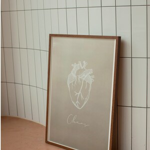 May include: A framed print with a white anatomical heart illustration and the word "Chaos" written in cursive below it. The print is on a light brown background and is framed in a dark brown wood frame.