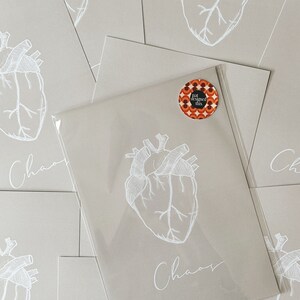 May include: A stack of beige cards with a white anatomical heart illustration and the word "Chaos" written in cursive. A small round sticker with the text "I designed this" is on the top card.