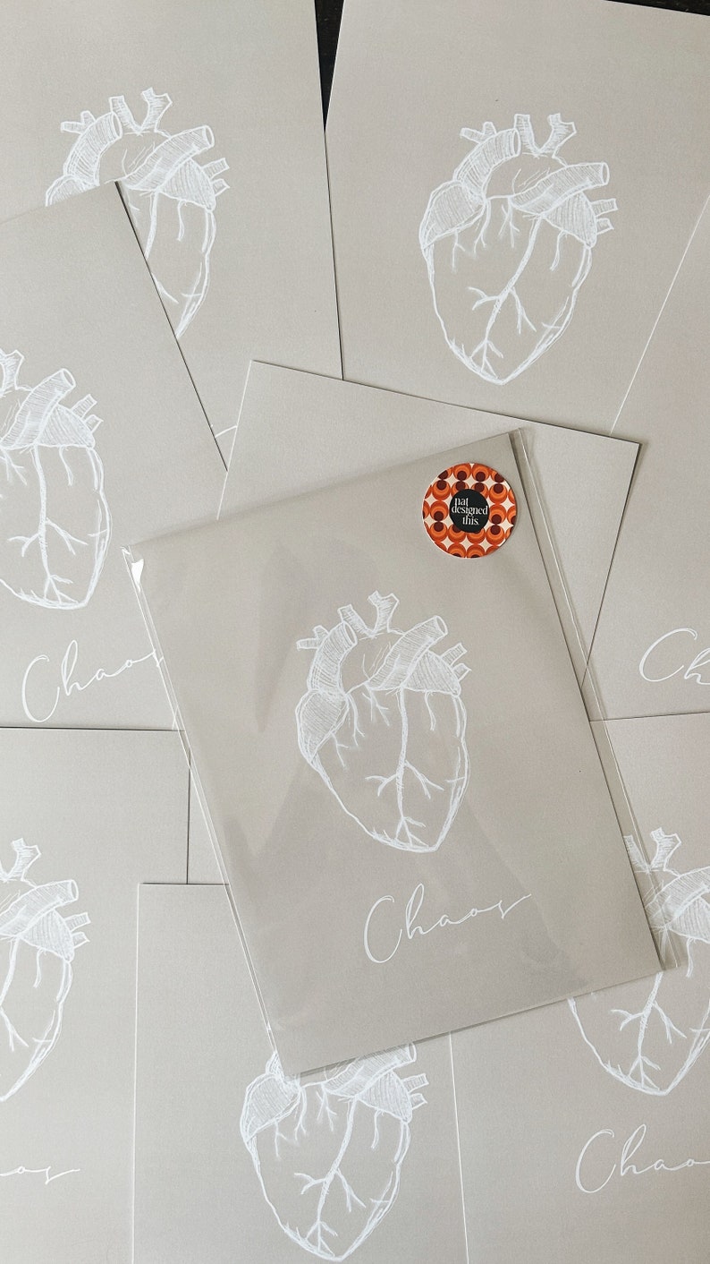May include: A stack of beige cards with a white anatomical heart illustration and the word "Chaos" written in cursive. A small round sticker with the text "I designed this" is on the top card.
