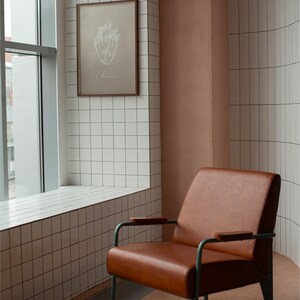 May include: A brown leather armchair with a metal frame and wooden armrests sits in a room with white tiled walls. A framed print of a heart with the word "Chaos" is hanging on the wall.