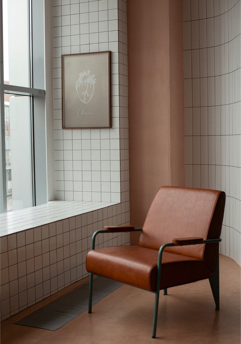 May include: A brown leather armchair with a metal frame and wooden armrests sits in a room with white tiled walls. A framed print of a heart with the word "Chaos" is hanging on the wall.