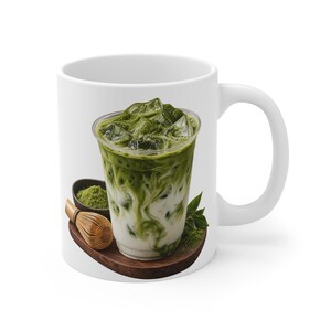 Könnte beinhalten: Ein weißer Keramikbecher mit einem aufgedruckten Motiv eines Eismatcha-Lattes in einem durchsichtigen Kunststoffbecher, ergänzt durch eine kleine Schale mit grünem Matchapulver, einen Bambusbesen und grüne Teeblätter auf einem Holzsockel.