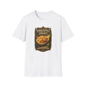 Puede incluir: Una camiseta blanca de manga corta con un estampado gráfico central de arroz frito con piña servido en una cáscara de piña. El diseño incluye el texto PINEAPPLE FRIED RICE, THAI STREET FOOD y AUTHENTIC THAI CUISINE dentro de un marco adornado.