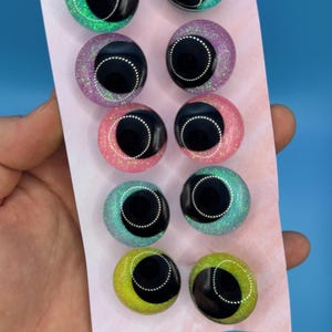 May include: Eight round, colourful, glittery googly eyes with black centres and white outlines. The eyes are arranged in two rows of four on a white card.