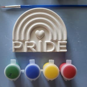 May include: A white, semi-circular plaster ornament featuring a raised rainbow design, a central heart, and the word PRIDE in block letters. The set includes a thin blue paintbrush and four small, round plastic pots containing green, blue, yellow, and red paint.