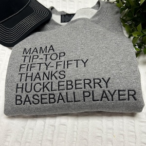 May include: A heather gray crewneck sweatshirt featuring black embroidered text that reads MAMA, TIP-TOP, FIFTY-FIFTY, THANKS, HUCKLEBERRY, BASEBALL PLAYER. The garment is paired with a black and white baseball cap positioned at the top left corner.