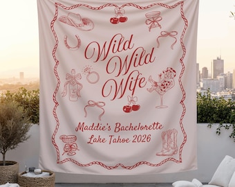 May include: A light pink banner with red text and illustrations, including a cowboy hat, cherries, and a horseshoe. The banner reads "Wild Wild Wife" and "Maddie's Bachelorette Lake Tahoe 2026". The banner is hanging outdoors.