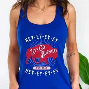 Op de afbeelding: Koningsblauw tanktop met de tekst "HEY-EY-EY-EY Let's Go BUFFALO HEY-EY-EY-EY" in het wit. Een rode buffelafbeelding is gecentreerd op de voorkant, met "EST. 1960" eronder. De tanktop heeft een racerback-ontwerp.