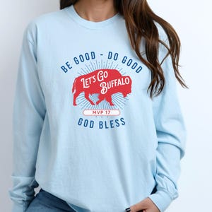 May include: Light blue long-sleeve shirt with the text "BE GOOD - DO GOOD" and "LET'S GO BUFFALO" in red. A red buffalo graphic is in the center, with "MVP 17" and "GOD BLESS" below. The shirt is made of a soft material.