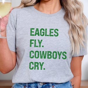 May include: A light gray t-shirt with the text "EAGLES FLY. COWBOYS CRY." in green block letters. The shirt is a casual style, suitable for everyday wear. The image shows the shirt being worn.