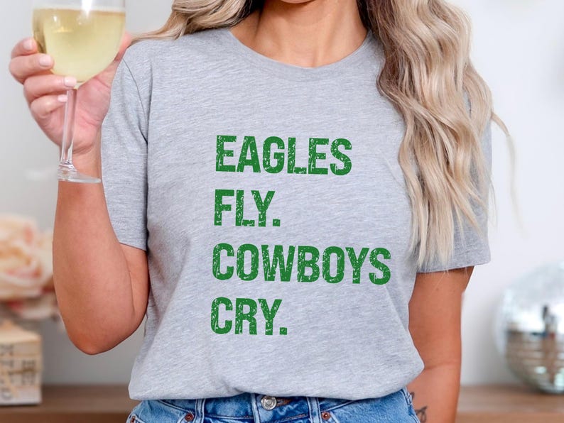 May include: A light gray t-shirt with the text "EAGLES FLY. COWBOYS CRY." in green block letters. The shirt is a casual style, suitable for everyday wear. The image shows the shirt being worn.