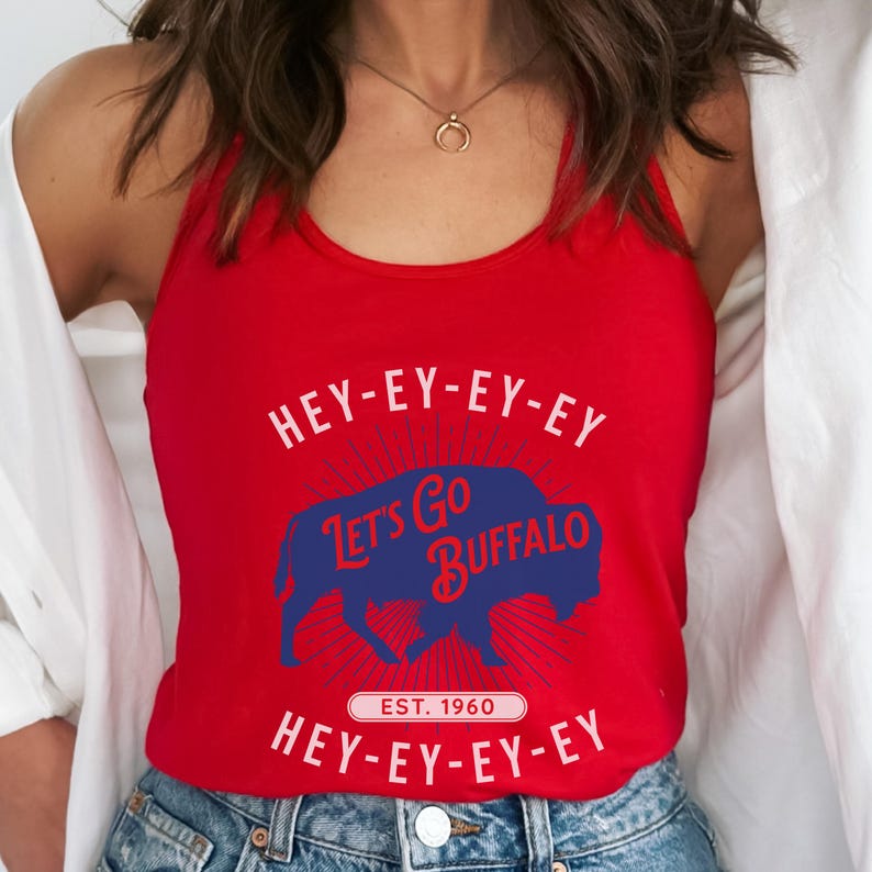 Op de afbeelding: Een rode tanktop met de tekst "HEY-EY-EY-EY, LET'S GO BUFFALO, EST. 1960, HEY-EY-EY-EY" in wit en blauw. Het ontwerp bevat een blauw buffelsilhouet. De top is mouwloos.