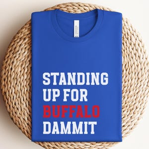 May include: A royal blue t-shirt with the words "STANDING UP FOR BUFFALO DAMMIT" in white and red block letters. The shirt is folded neatly on a woven, circular placemat.