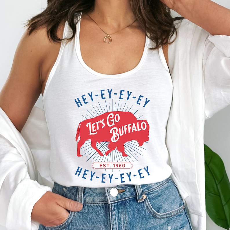 Op de afbeelding: Witte tanktop met een rode buffelafbeelding en de tekst "Let's Go Buffalo". Het ontwerp bevat de tekst "HEY-EY-EY-EY" en "EST. 1960". De tanktop is een casual stijl.