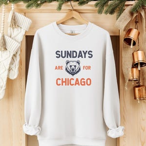 K&ouml;nnte beinhalten: Wei&szlig;er Pullover mit dem Text "Sundays are for Chicago" in Marineblau und Orange. Eine B&auml;renkopfgrafik befindet sich in der Mitte des Textes.