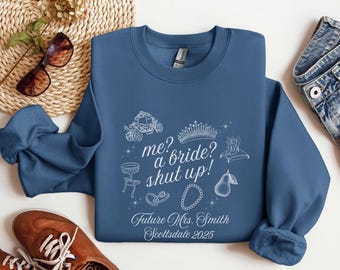 Op de afbeelding: Een blauwe sweatshirt met een witte grafische afbeelding met een koets, kroon en andere bruidsgerelateerde illustraties. De tekst luidt "me? a bride? shut up!" en "Future Mrs. Smith Scottsdale 2025".