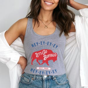 Op de afbeelding: Een grijze tanktop met de tekst "HEY-EY-EY-EY, LET'S GO BUFFALO, HEY-EY-EY-EY" in blauw en rood. Het ontwerp bevat een rood buffelsilhouet en de tekst "EST. 1960".