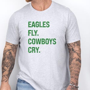 May include: Light gray t-shirt with the text "EAGLES FLY. COWBOYS CRY." in green block letters. The shirt is short-sleeved and has a crew neck. The text is centered on the front of the shirt.
