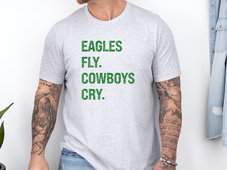 May include: Light gray t-shirt with the text "EAGLES FLY. COWBOYS CRY." in green block letters. The shirt is short-sleeved and has a crew neck. The text is centered on the front of the shirt.