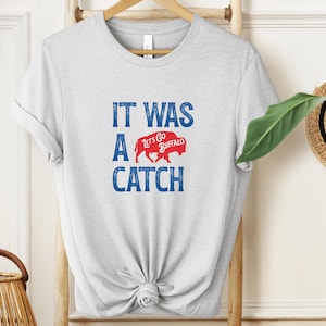 May include: Light gray t-shirt with the text "IT WAS A CATCH" in blue, and a red buffalo graphic with "Lets Go Buffalo" text. The shirt is hanging on a wooden ladder.