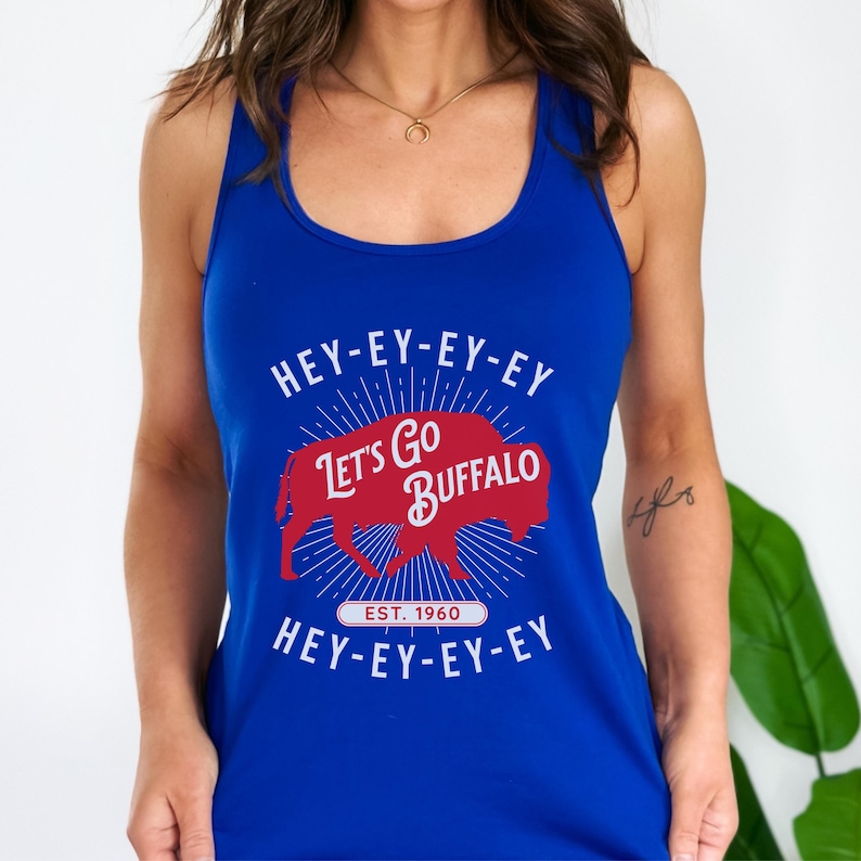 Op de afbeelding: Koningsblauw tanktop met de tekst "HEY-EY-EY-EY Let's Go BUFFALO HEY-EY-EY-EY" in het wit. Een rode buffelafbeelding is gecentreerd op de voorkant, met "EST. 1960" eronder. De tanktop heeft een racerback-ontwerp.