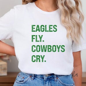 May include: White t-shirt with the text "EAGLES FLY. COWBOYS CRY." in green, block letters. The shirt is made of a soft material and is suitable for casual wear. The shirt is paired with blue jeans.