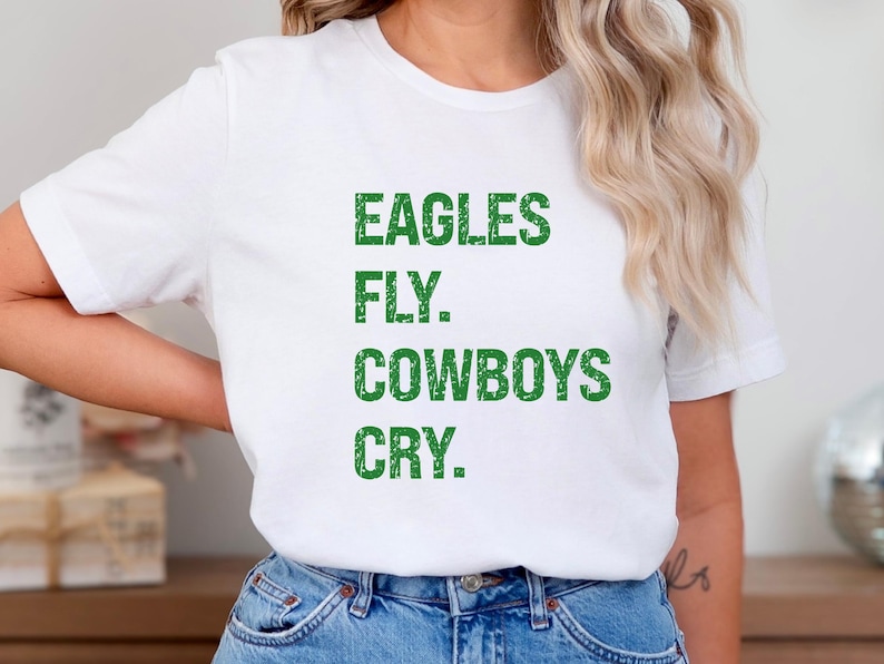 May include: White t-shirt with the text "EAGLES FLY. COWBOYS CRY." in green, block letters. The shirt is made of a soft material and is suitable for casual wear. The shirt is paired with blue jeans.