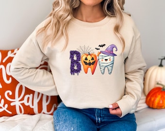 May include: Beige sweatshirt with a Halloween-themed graphic. The graphic spells "BOO" with a purple letter B, an orange jack-o'-lantern, and a tooth wrapped in bandages wearing a witch hat. Black bats and a spiderweb are also included.