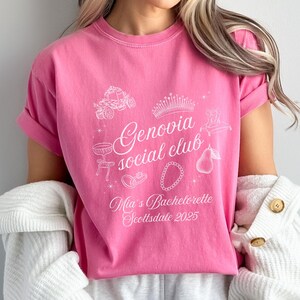 May include: Pink t-shirt with white graphic text and illustrations. The text reads "Genovia social club" with "Mia's Bachelorette Scottsdale 2025" below. Illustrations include a crown, carriage, and jewelry.