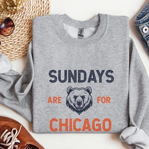 K&ouml;nnte beinhalten: Ein grauer Pullover mit dem Text "Sundays are for Chicago" in Marineblau und Orange. Eine B&auml;rengrafik befindet sich in der Mitte des Textes.