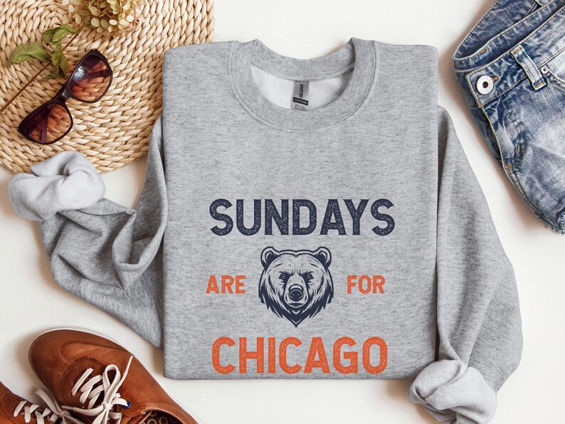 K&ouml;nnte beinhalten: Ein grauer Pullover mit dem Text "Sundays are for Chicago" in Marineblau und Orange. Eine B&auml;rengrafik befindet sich in der Mitte des Textes.