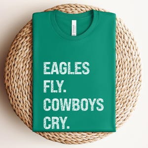 May include: A folded, emerald green t-shirt with the text "EAGLES FLY. COWBOYS CRY." in white, block letters. The shirt is resting on a round, woven wicker placemat.