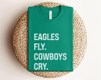 May include: A folded, emerald green t-shirt with the text "EAGLES FLY. COWBOYS CRY." in white, block letters. The shirt is resting on a round, woven wicker placemat.