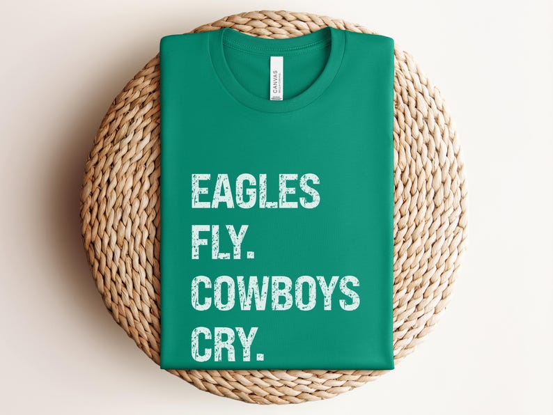 May include: A folded, emerald green t-shirt with the text "EAGLES FLY. COWBOYS CRY." in white, block letters. The shirt is resting on a round, woven wicker placemat.