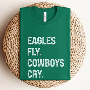 May include: A folded green t-shirt with the text "EAGLES FLY. COWBOYS CRY." in distressed white lettering. The shirt is displayed on a round, woven wicker placemat.