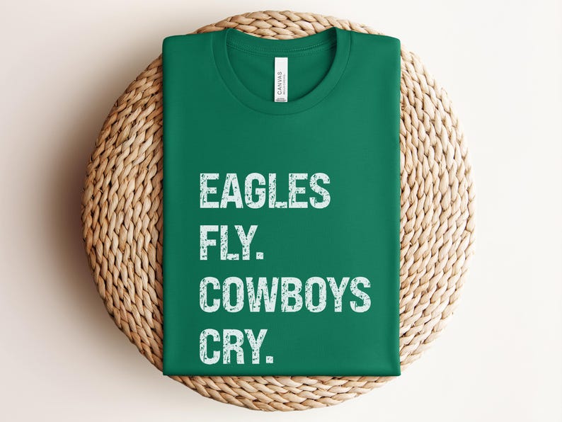 May include: A folded green t-shirt with the text "EAGLES FLY. COWBOYS CRY." in distressed white lettering. The shirt is displayed on a round, woven wicker placemat.