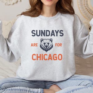 K&ouml;nnte beinhalten: Ein grauer Pullover mit dem Text "Sundays are for Chicago" in Marineblau und Orange. Eine B&auml;rengrafik befindet sich mittig &uuml;ber dem Text.