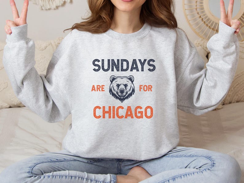 K&ouml;nnte beinhalten: Ein grauer Pullover mit dem Text "Sundays are for Chicago" in Marineblau und Orange. Eine B&auml;rengrafik befindet sich mittig &uuml;ber dem Text.