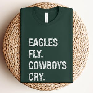 May include: Dark green t-shirt with the text "EAGLES FLY. COWBOYS CRY." in white, displayed on a woven, circular placemat. The shirt has a crew neck and a tag at the neckline.