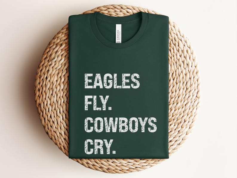 May include: Dark green t-shirt with the text "EAGLES FLY. COWBOYS CRY." in white, displayed on a woven, circular placemat. The shirt has a crew neck and a tag at the neckline.