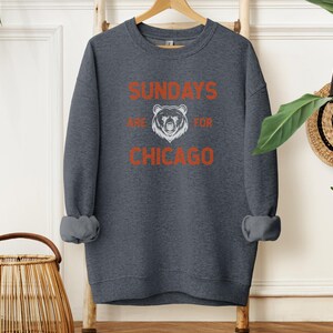 K&ouml;nnte beinhalten: Ein dunkelgrauer Pullover mit dem Text "Sundays are for Chicago" in Orange und Wei&szlig;. Eine wei&szlig;e B&auml;rensilhouette befindet sich in der Mitte des Textes.