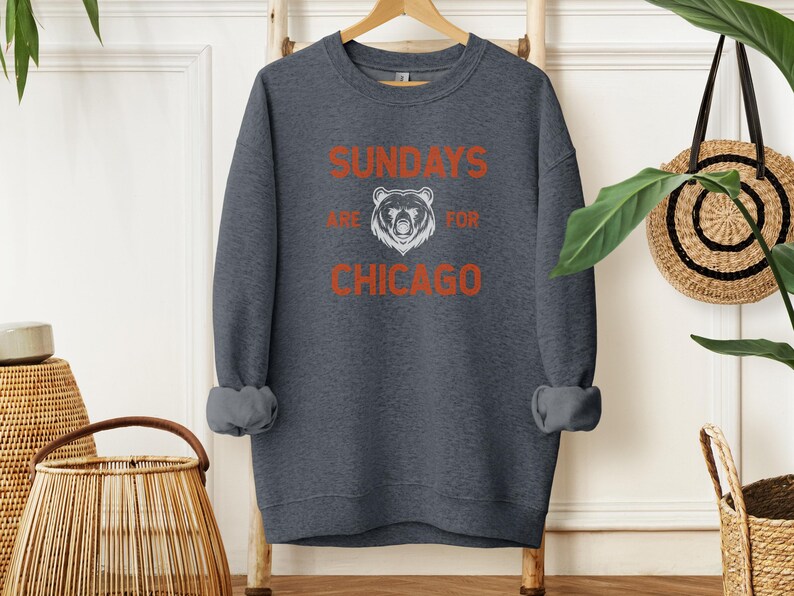 K&ouml;nnte beinhalten: Ein dunkelgrauer Pullover mit dem Text "Sundays are for Chicago" in Orange und Wei&szlig;. Eine wei&szlig;e B&auml;rensilhouette befindet sich in der Mitte des Textes.