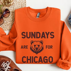 K&ouml;nnte beinhalten: Orangefarbener Pullover mit einem schwarzen grafischen Aufdruck, der "Sundays are for Chicago" mit einer B&auml;renillustration sagt.